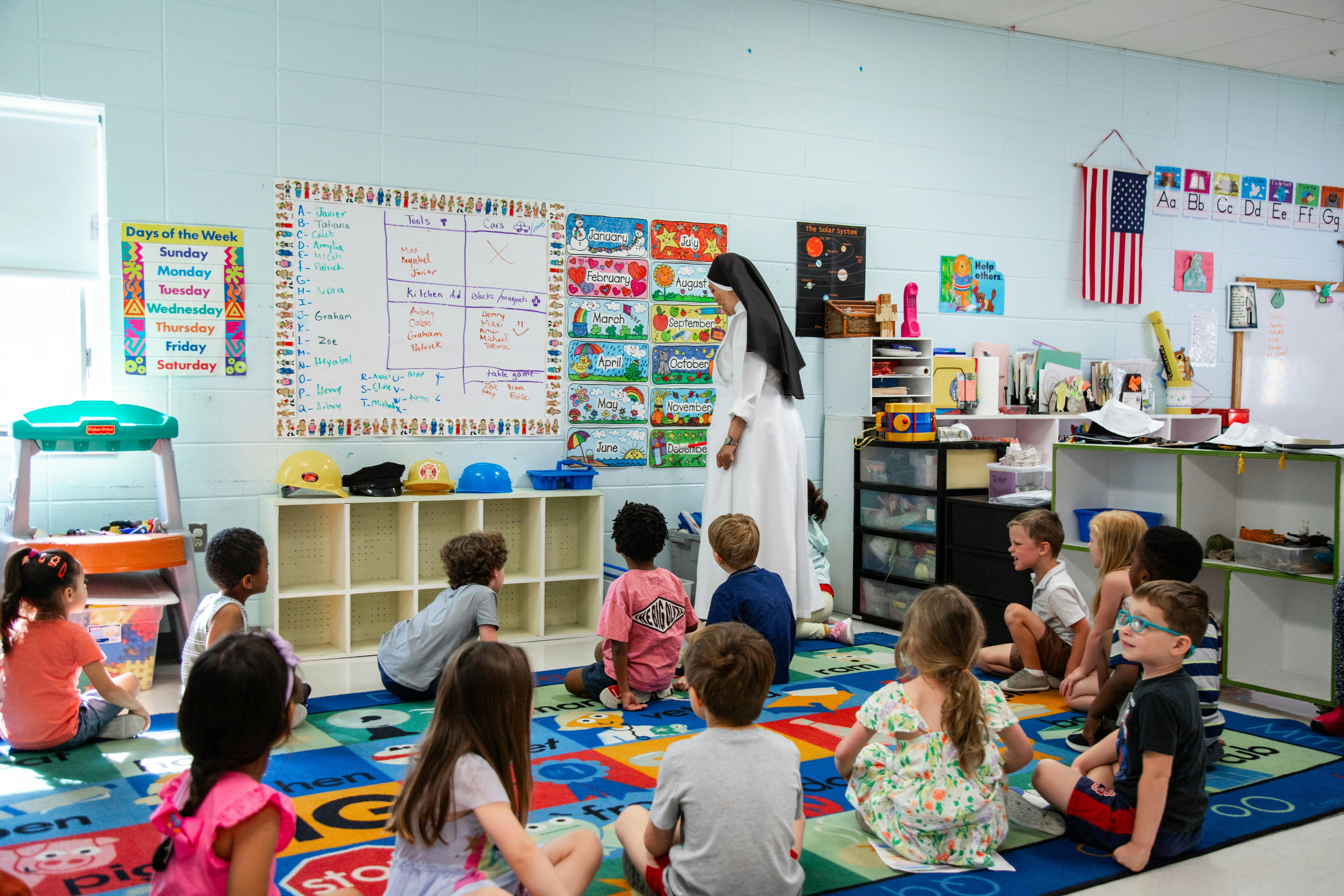 Person in white dress teaching children sitting on a colorful rug in a classroom