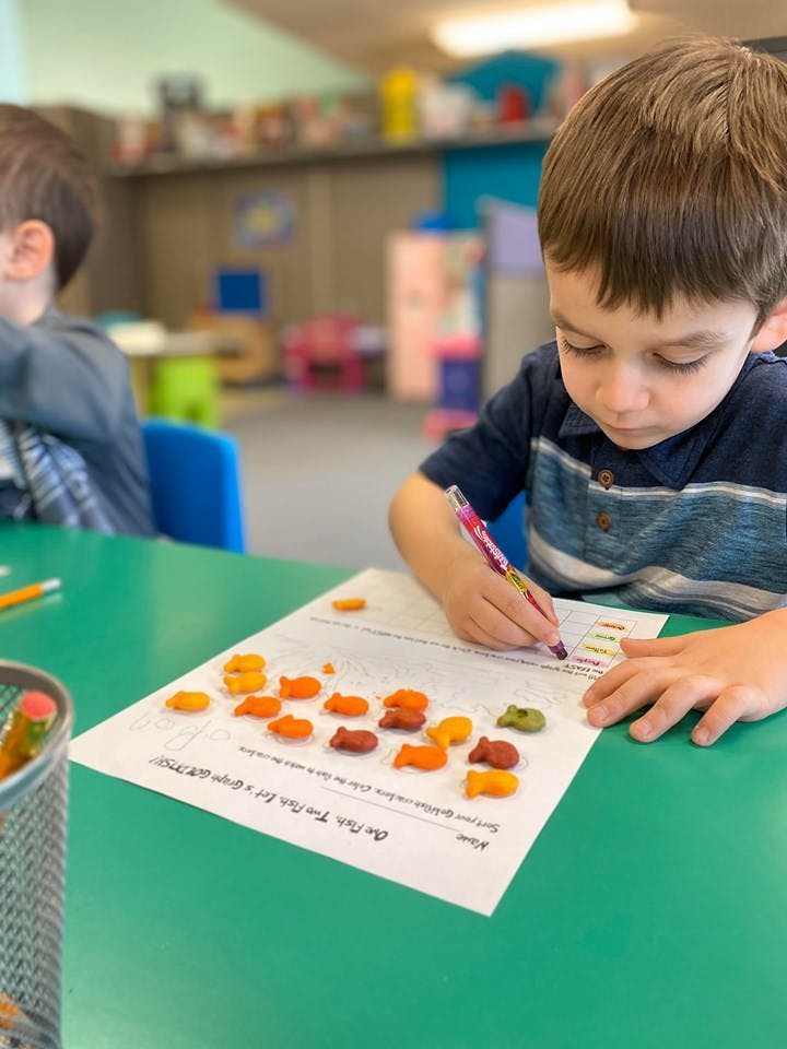 Child writing on paper with fish-shaped crackers on a table in a classroom