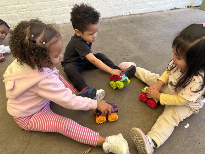 Three children sitting on the ground playing with toy monster trucks