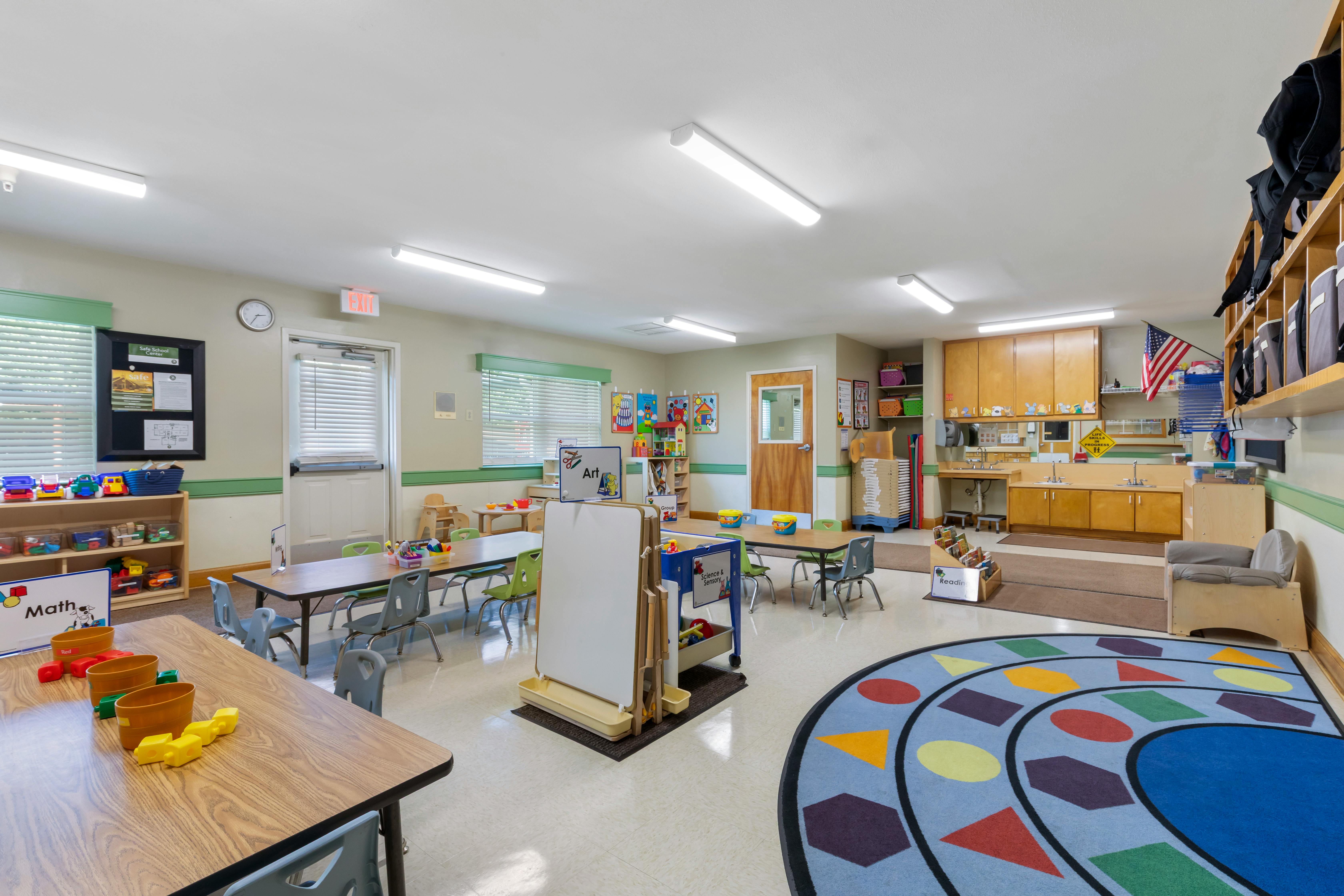 Classroom with tables, chairs, colorful rug, shelves with toys, and sinks
