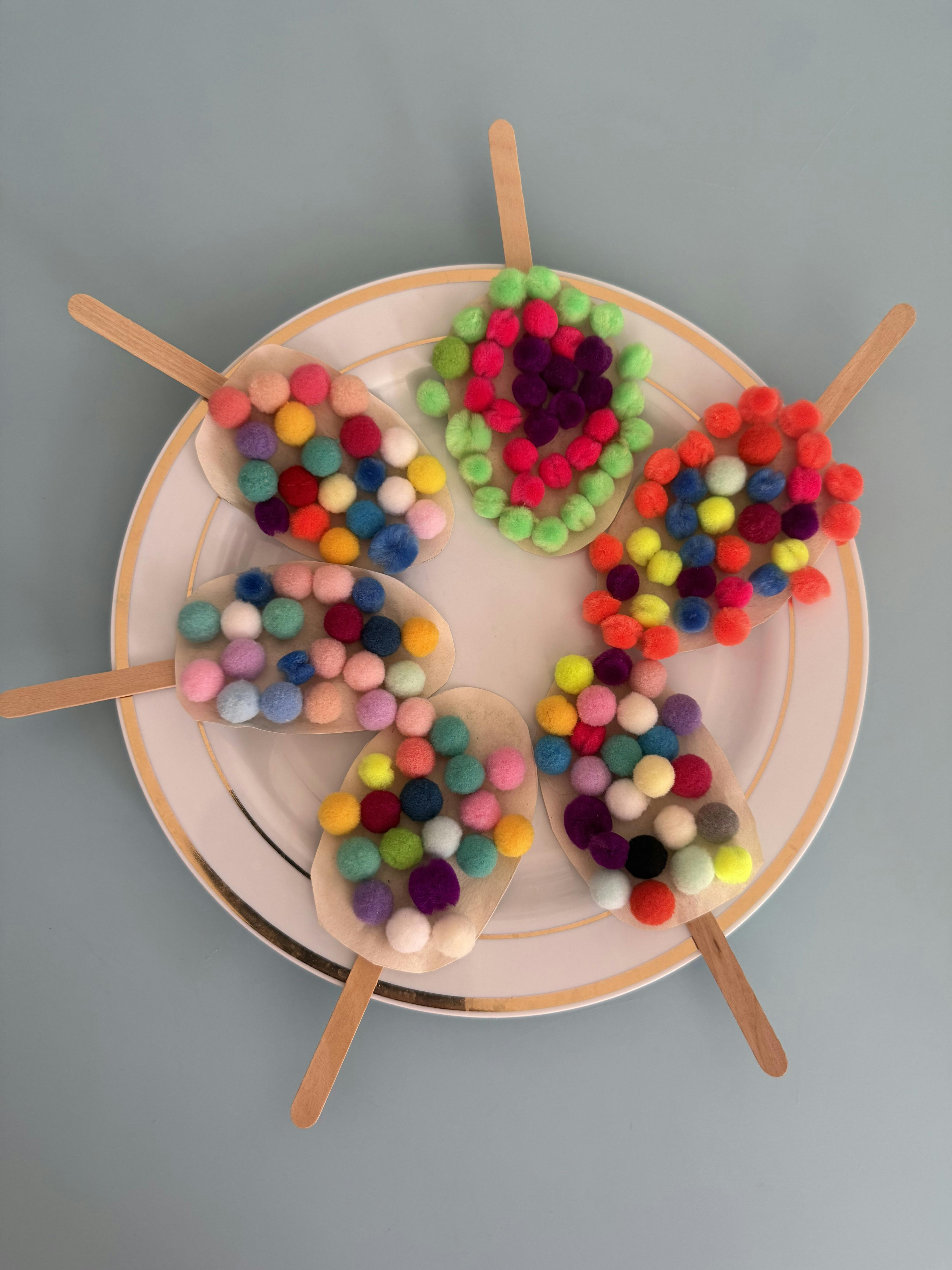 Craft popsicle shapes made of paper and colorful pom-poms on a plate