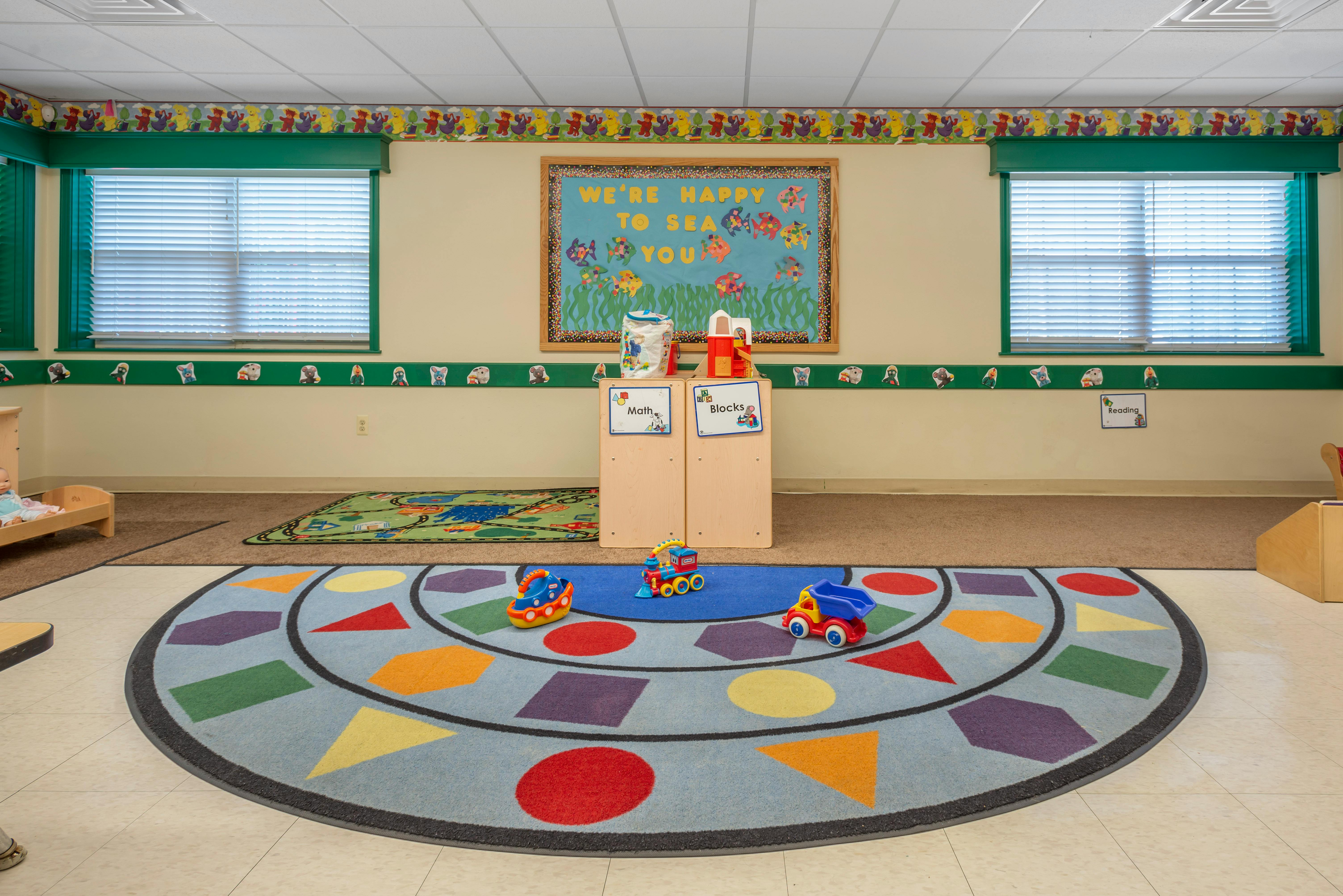 Classroom with colorful rug, toys, and a bulletin board that reads "We're Happy to Sea You"
