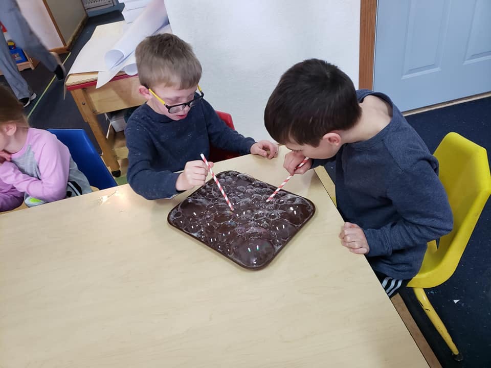 Two children blowing through straws into a tray of brown material