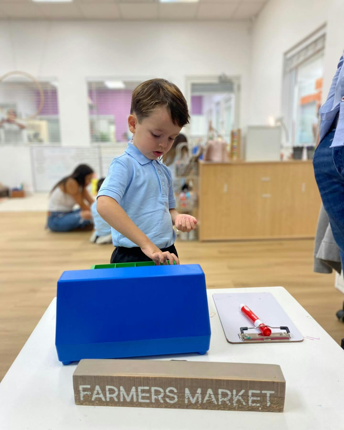 Child standing at a table with a blue box and a sign that says "FARMERS MARKET"