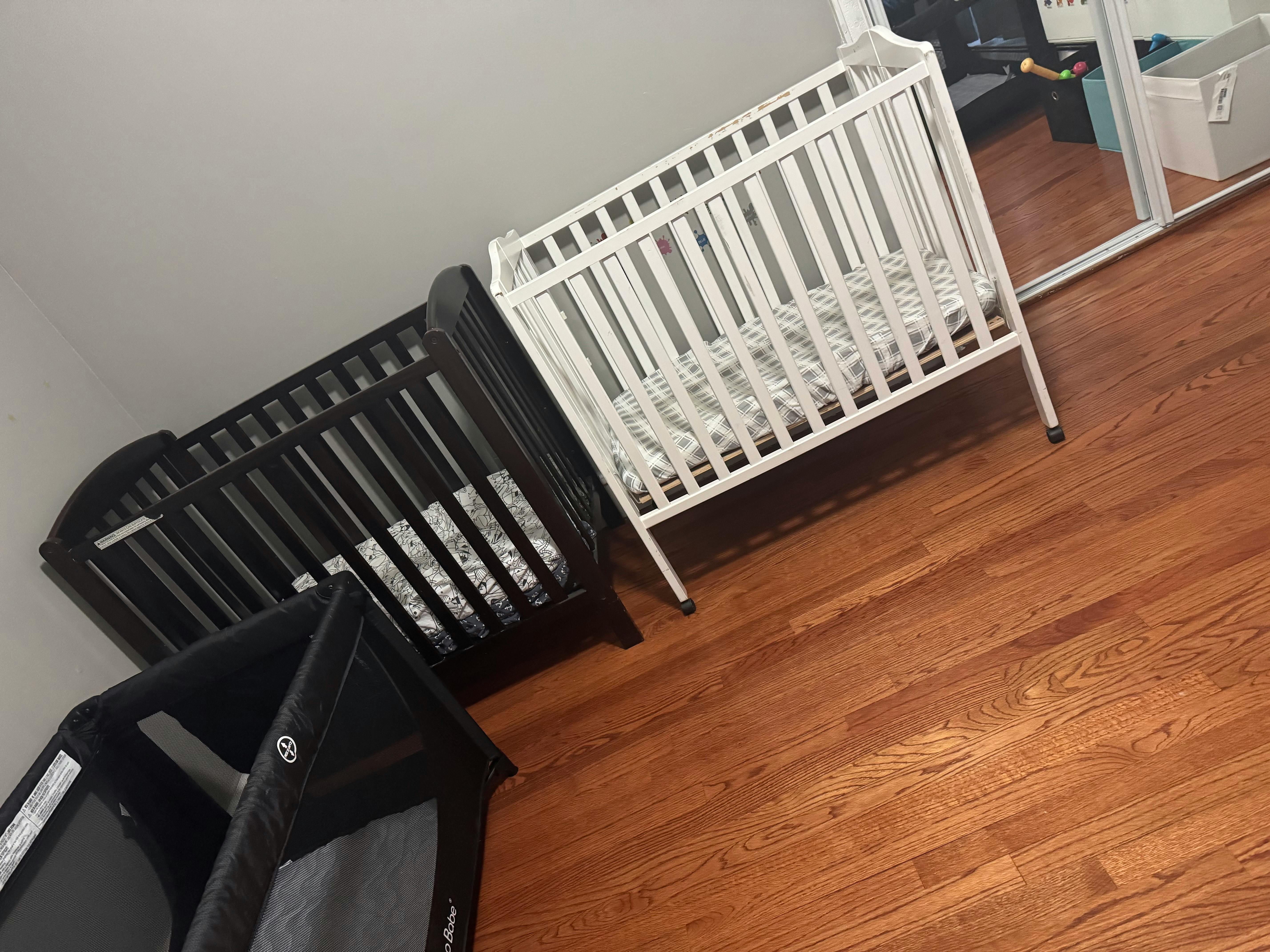 Three cribs, two wooden and one portable, on a wooden floor