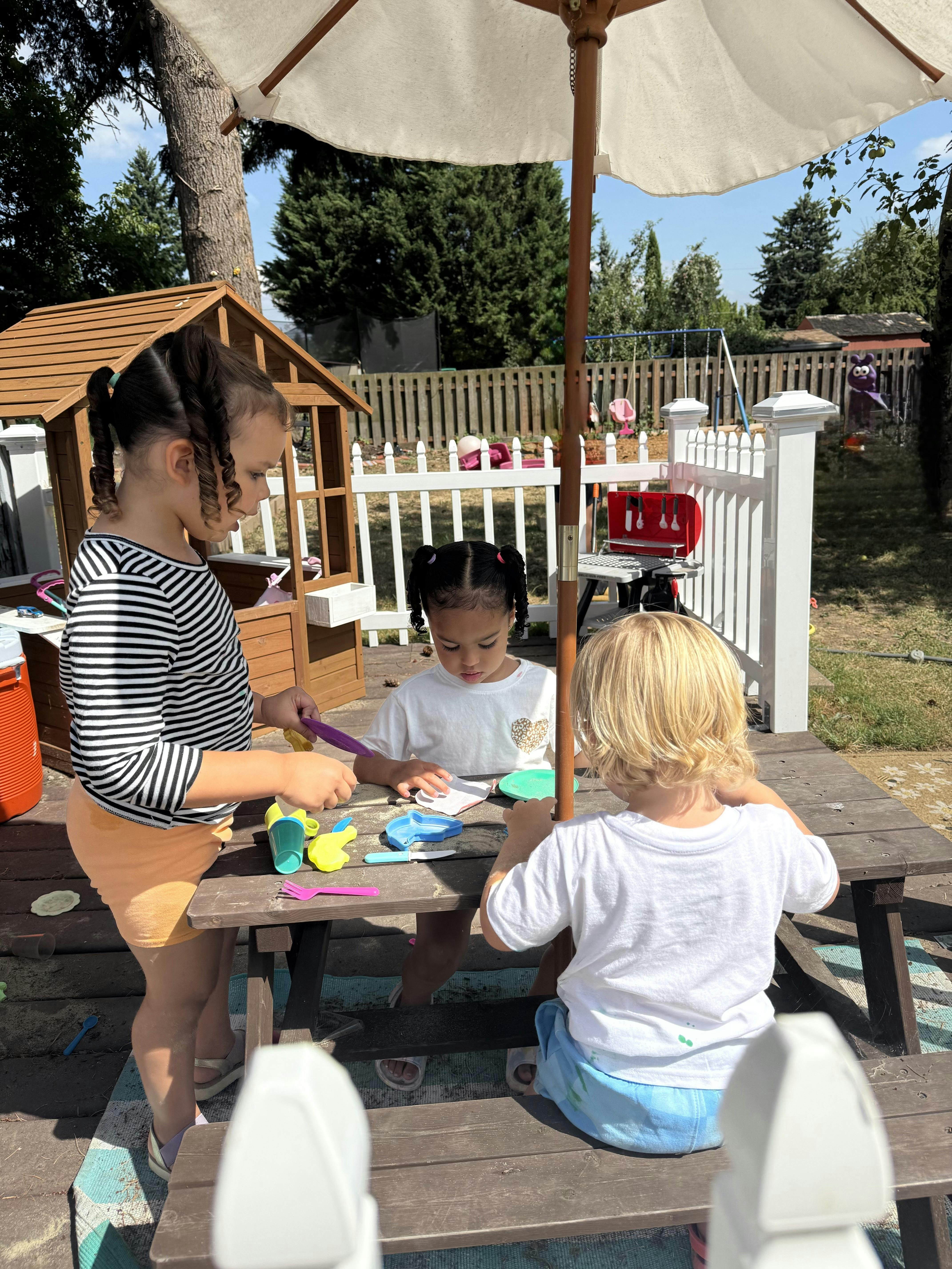 Three children playing with plastic toy dishes at a picnic table outdoors