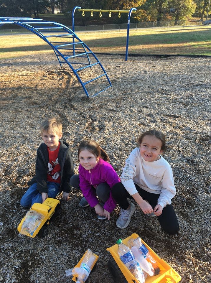 Three children crouching on wood chips with plastic bottles in toy trucks