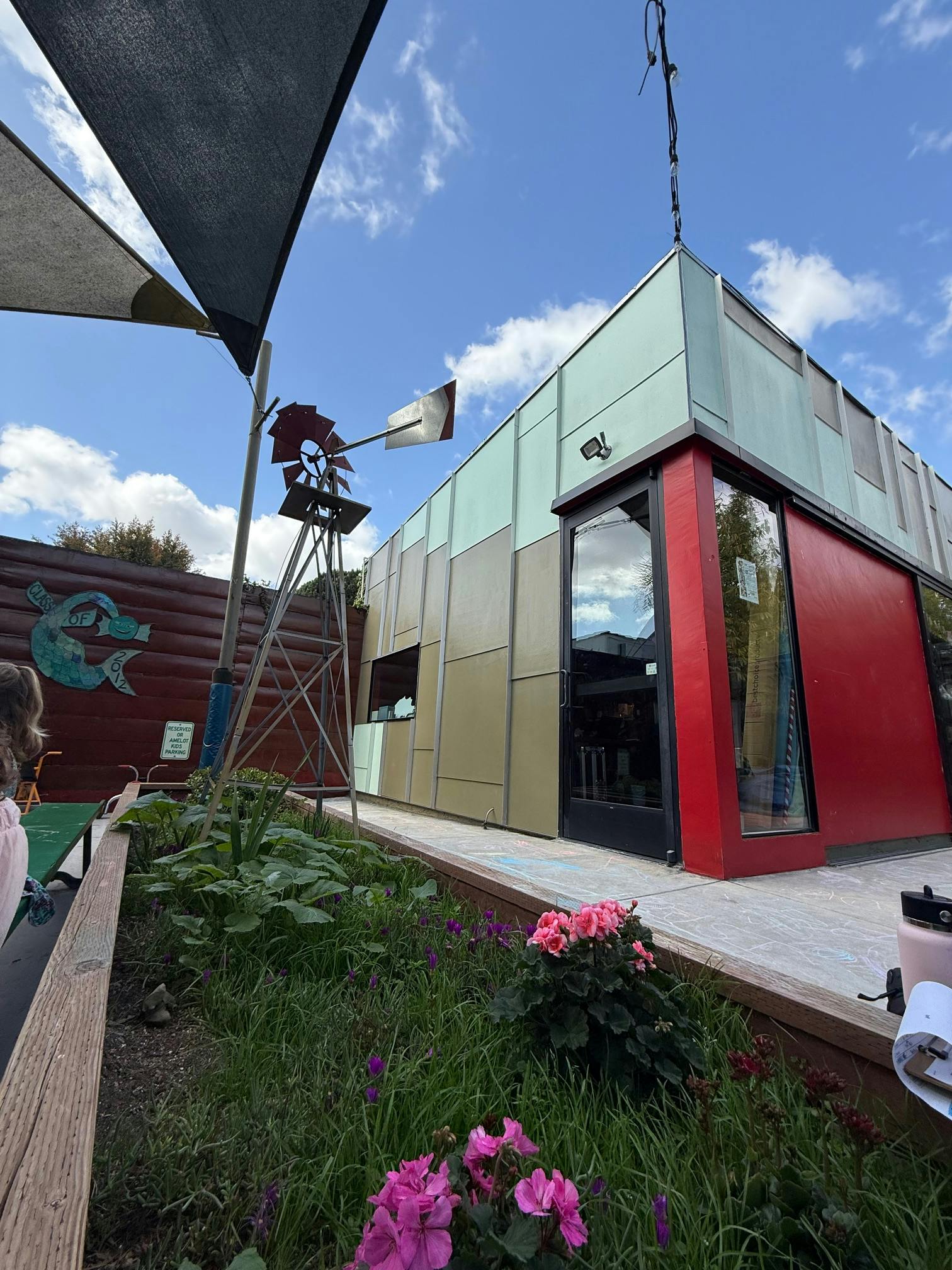 Building with red and green panels, a windmill, and plants in the foreground