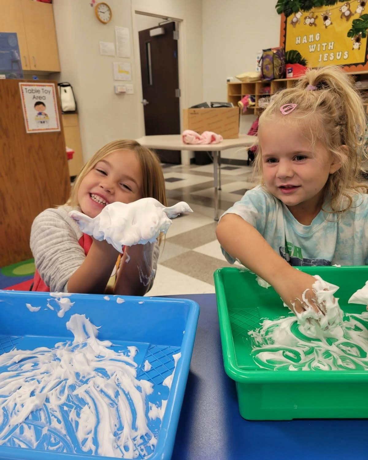 Two children playing with shaving cream in trays in a classroom