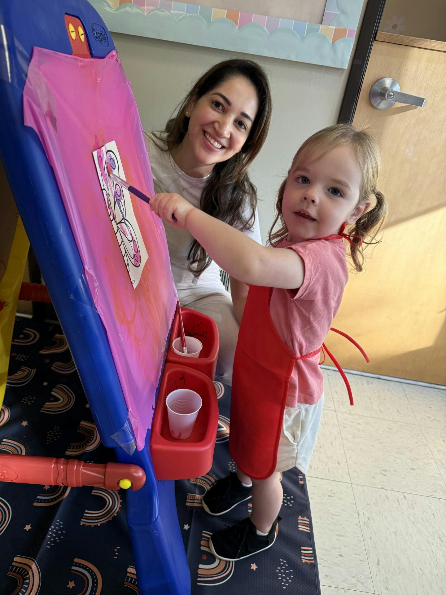 A person and a child painting on an easel in a classroom