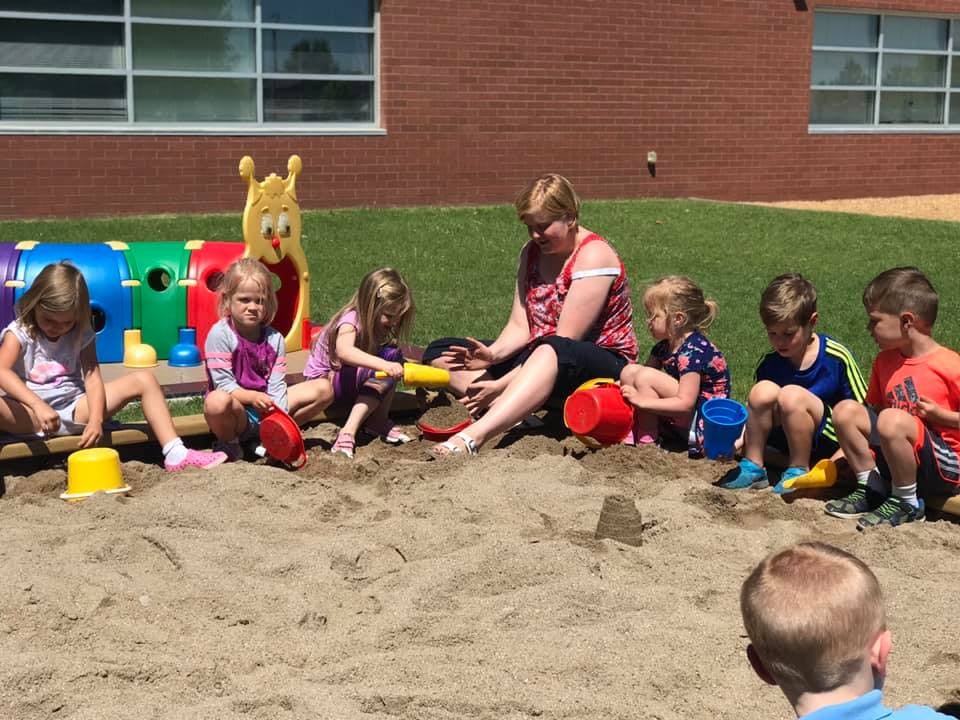 Adult and multiple children playing in a sandbox with toys