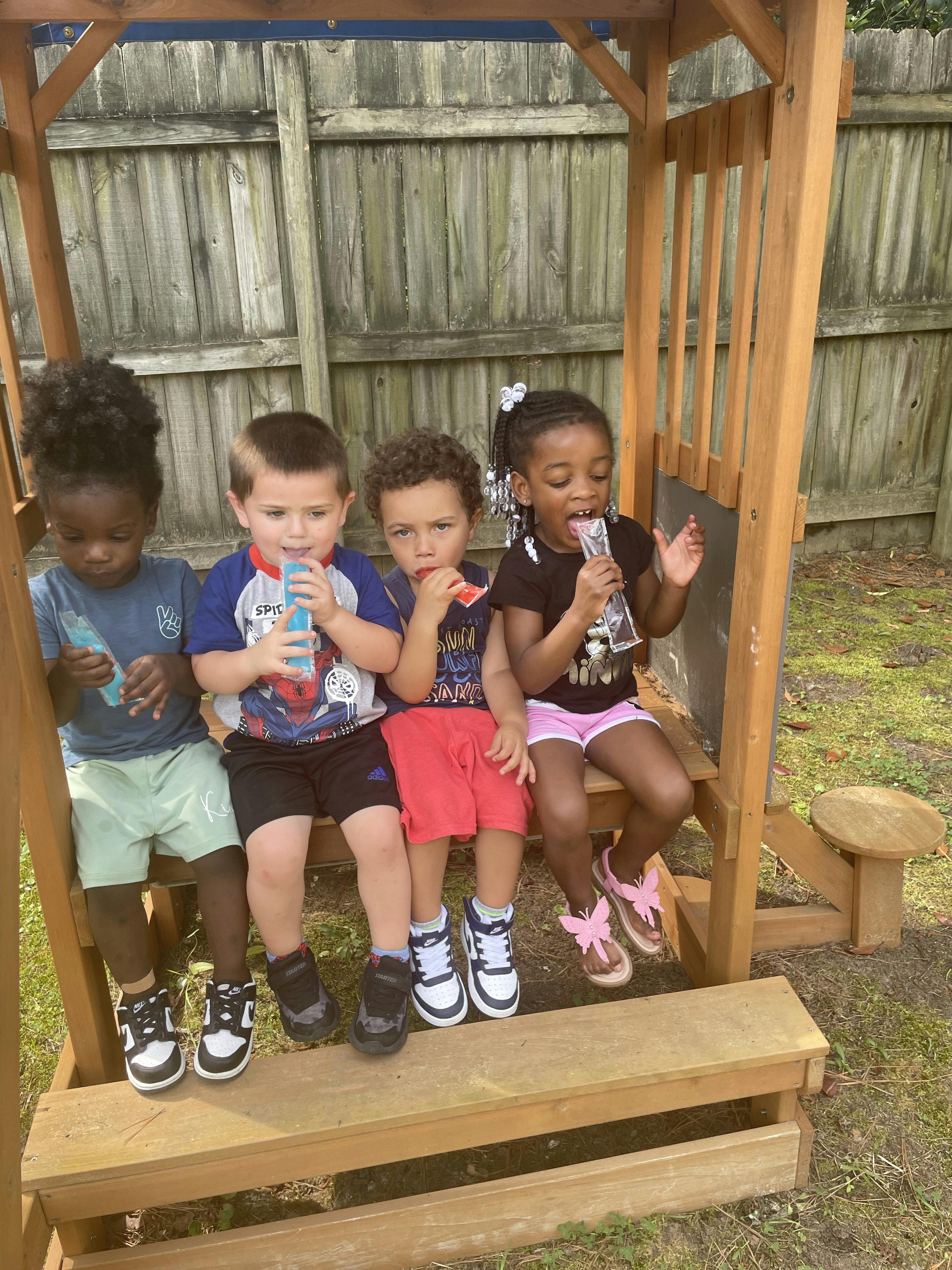 Four children sitting on a wooden play structure eating popsicles