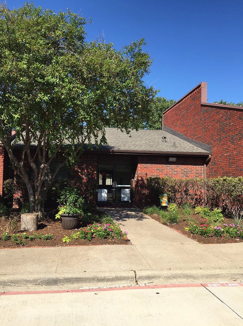 Brick building with a walkway leading to the entrance, surrounded by trees and plants