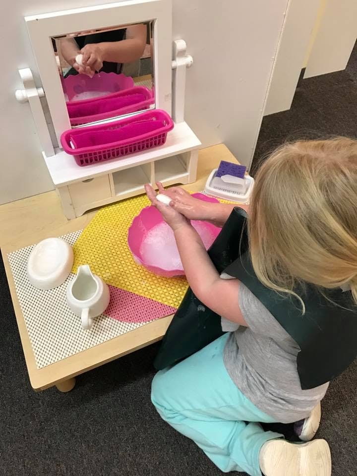 Child washing hands in a pink bowl with a mirror in front