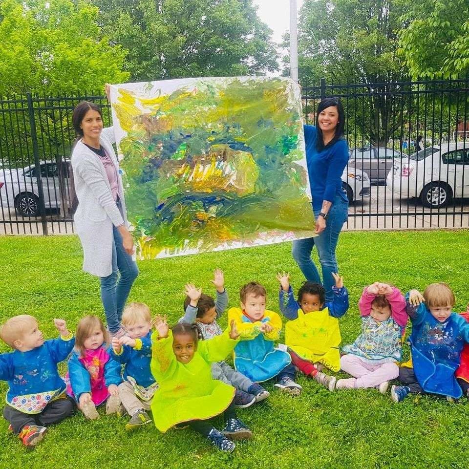 Two people holding up a large painting with children sitting on the grass below