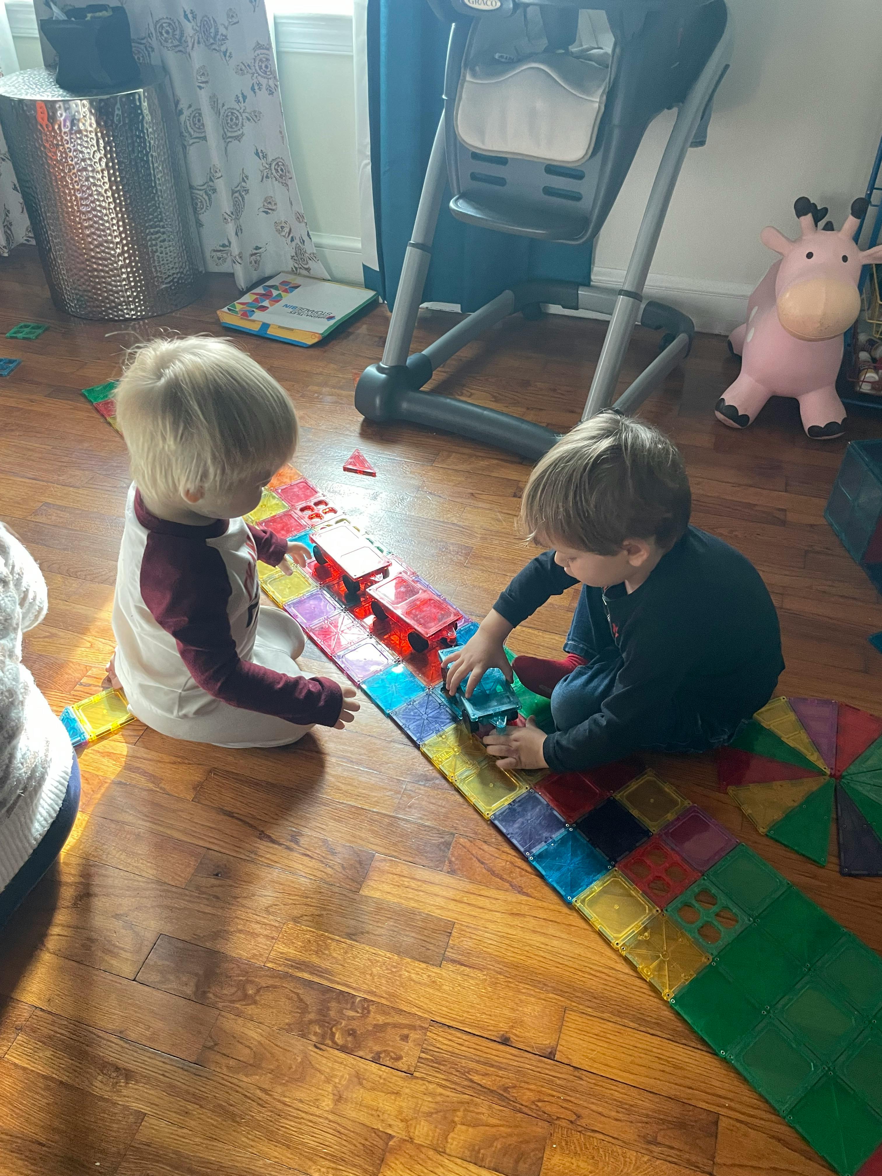 Two children playing with colorful magnetic tiles on a wooden floor