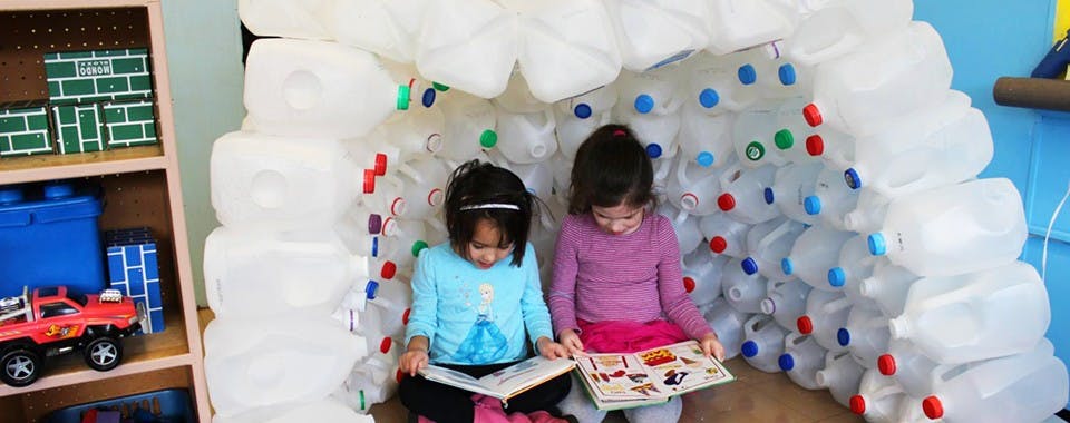 Two children reading books inside a structure made of plastic bottles