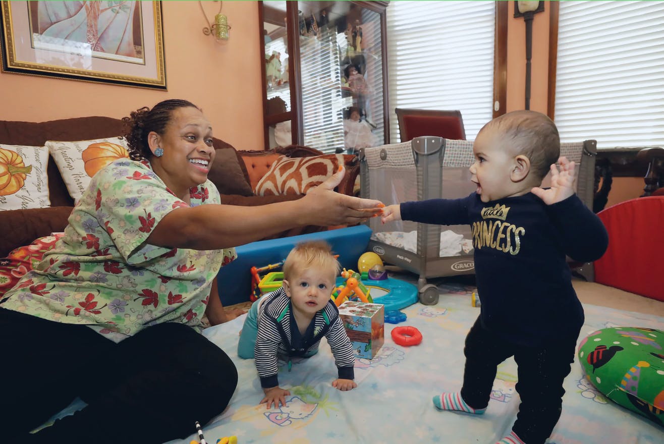 Adult reaching out to a toddler while an infant crawls on a play mat