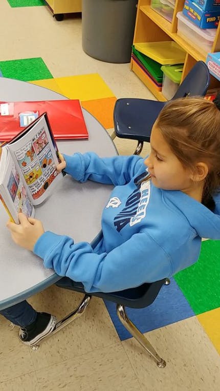 Child in blue sweatshirt reading a book at a table in a classroom
