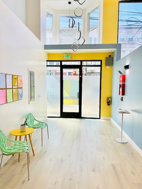 Waiting area with two green chairs and a yellow table, facing a glass door