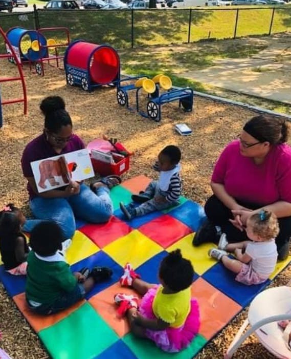 Adult reading a book to children on a colorful mat outdoors