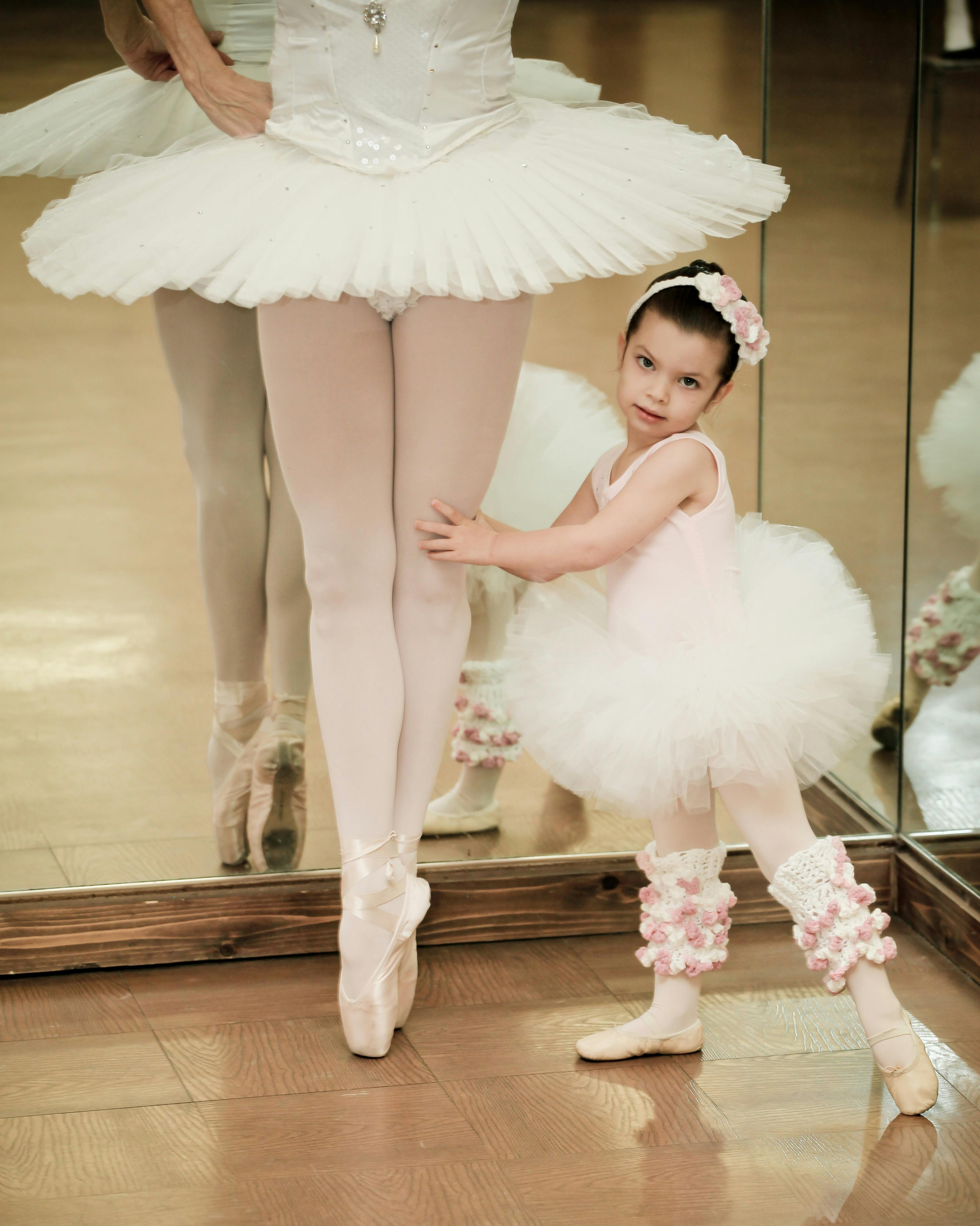 Child in a tutu holding onto a person in a tutu and pointe shoes