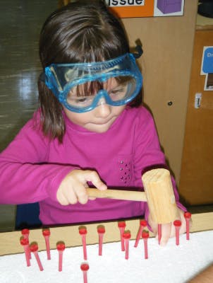 Child wearing safety goggles hammering red pegs into a board