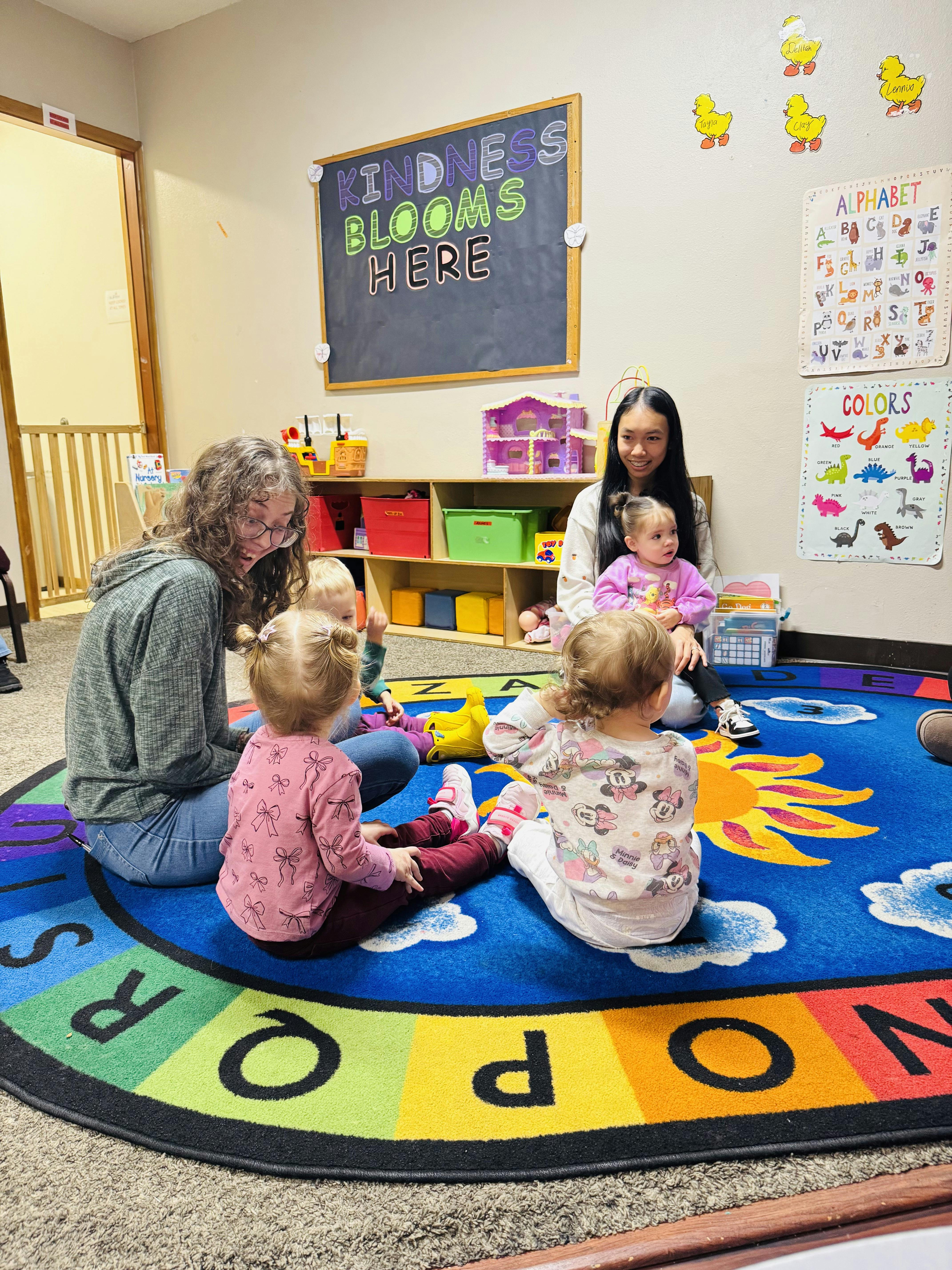 Two adults and four children sitting on a colorful alphabet rug in a classroom