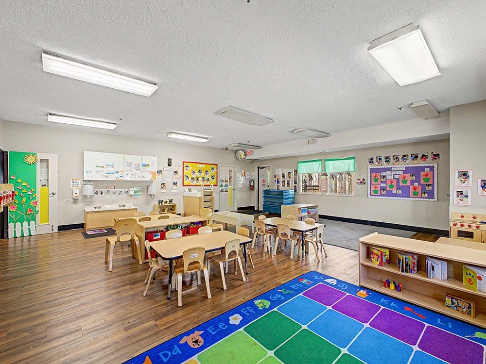 Empty classroom with small tables and chairs, colorful rug, and bulletin boards