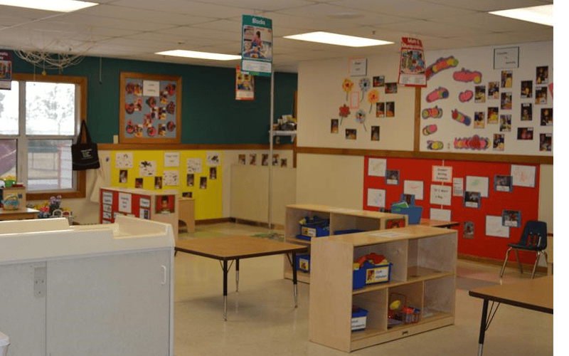 Classroom with tables, chairs, shelving units, and children's artwork on walls