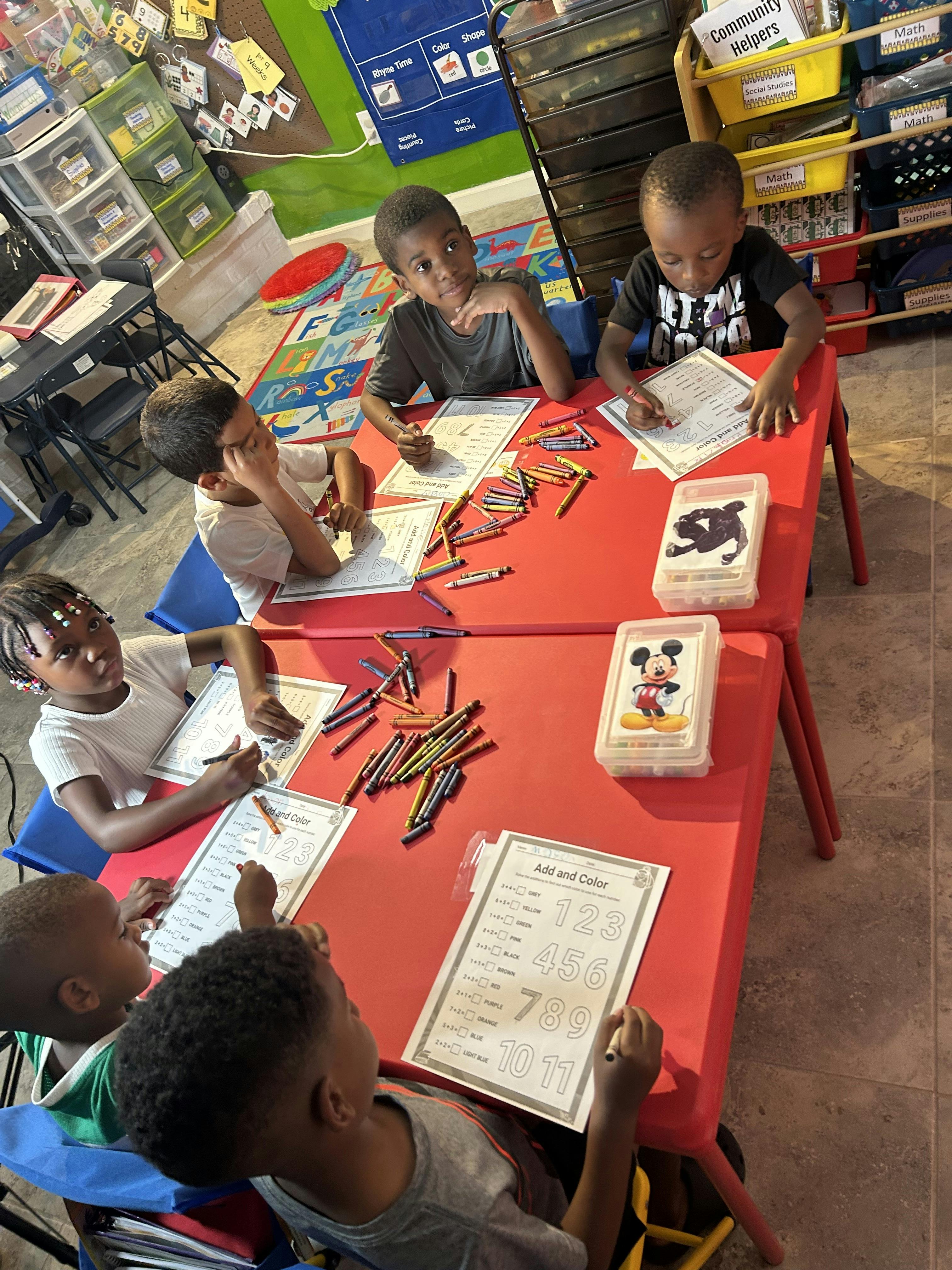 Children sitting at red tables, coloring and writing on papers in a classroom