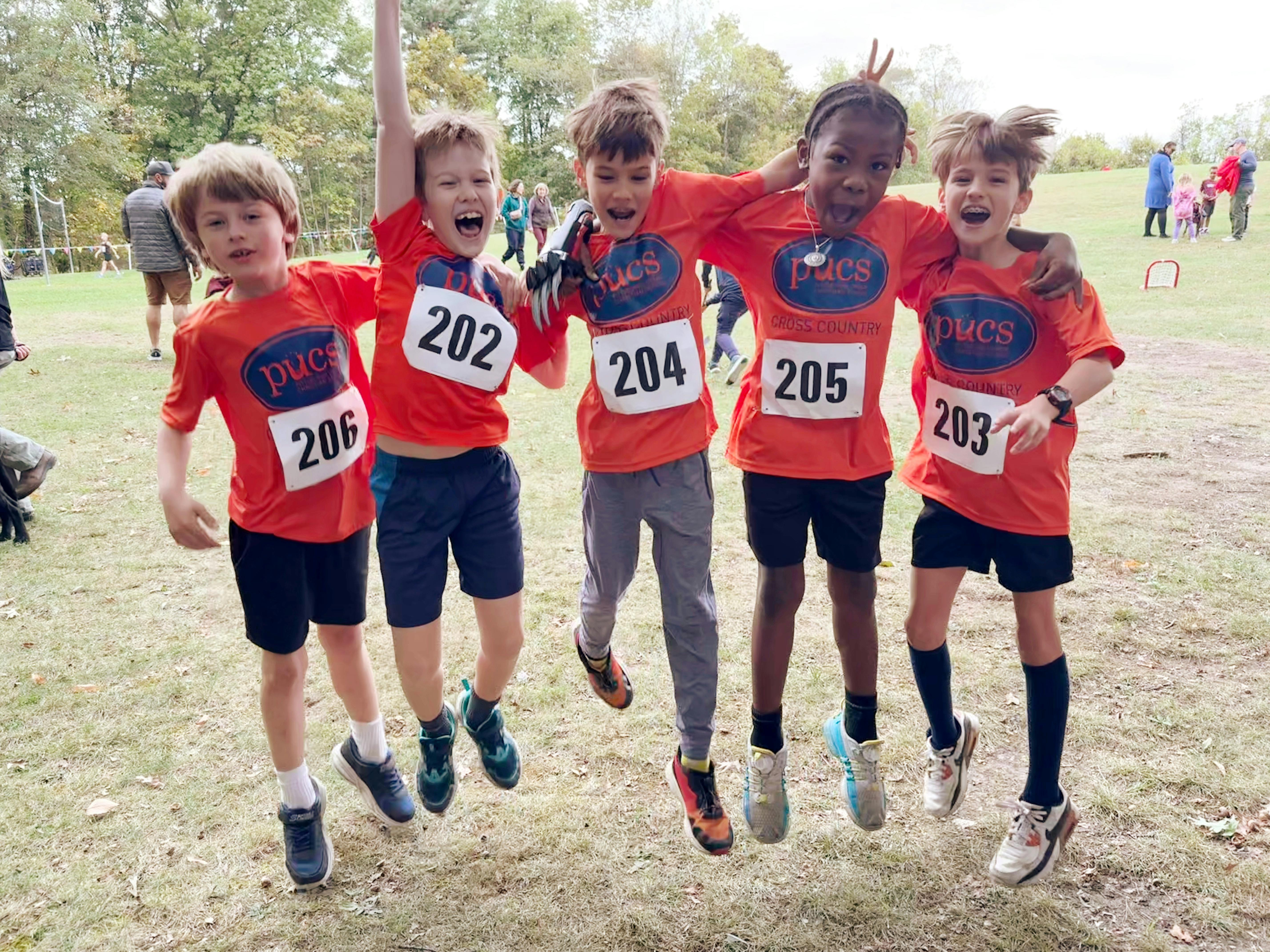 Five children wearing orange cross-country shirts with bib numbers jumping