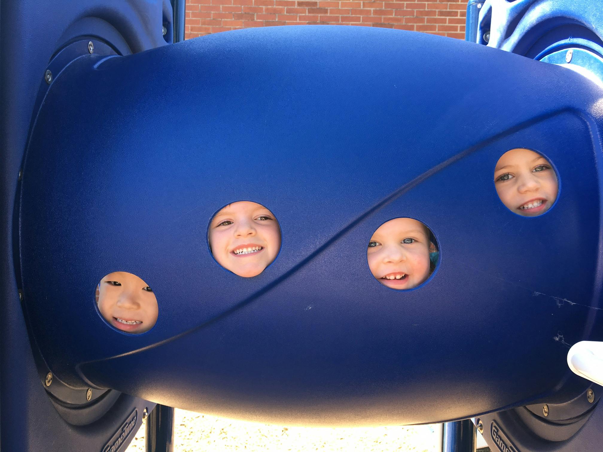 Three children looking through holes in a blue playground tunnel