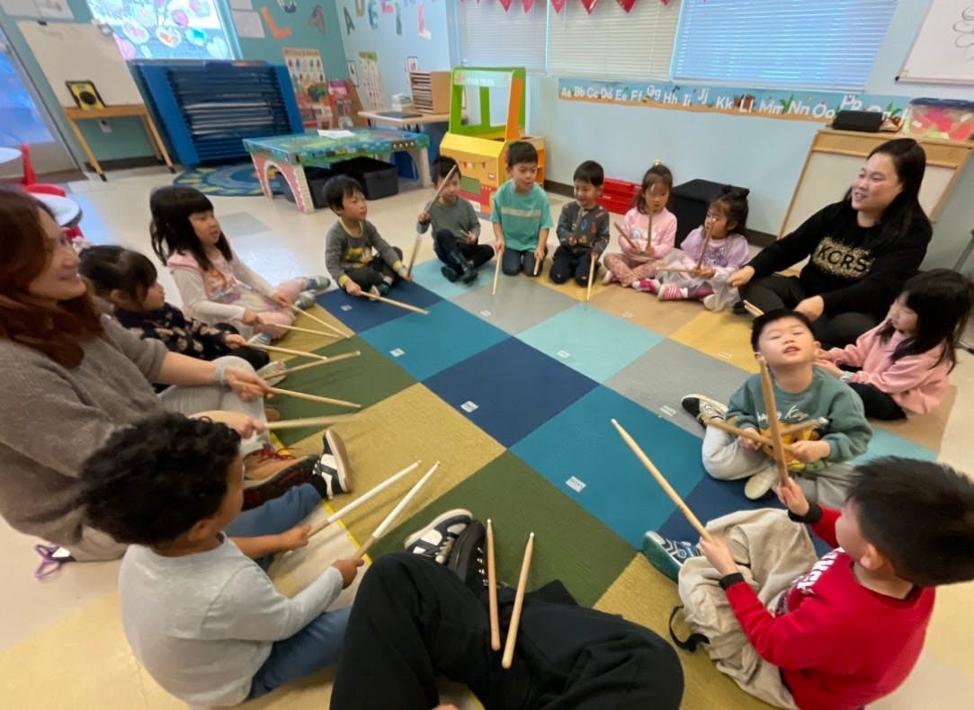 Adults and children sitting in a circle on the floor with drumsticks