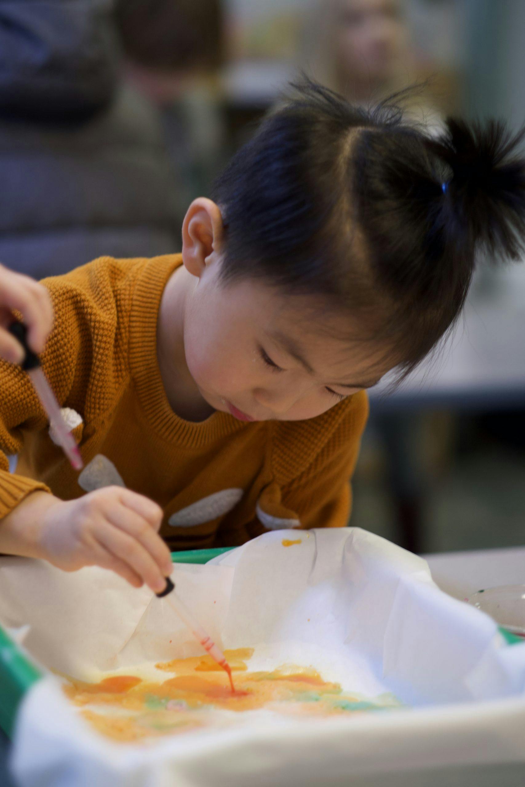 Child in orange sweater using a dropper to add liquid to paper
