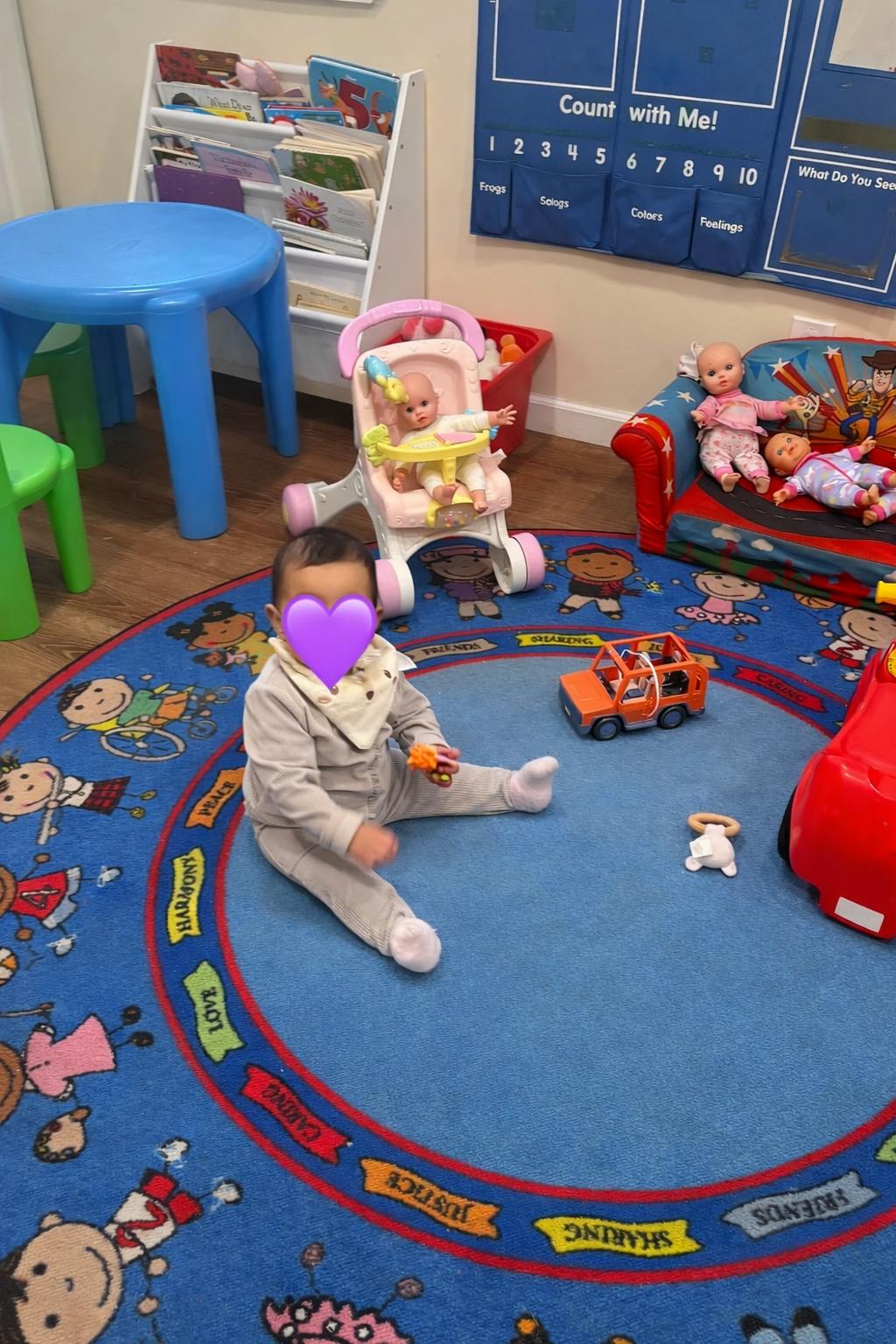 Child sitting on a blue rug with toys, dolls, and child-sized furniture