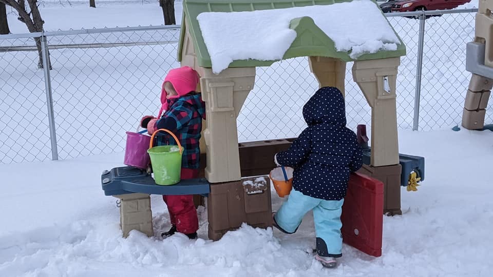 Two children playing with buckets in the snow next to a playhouse