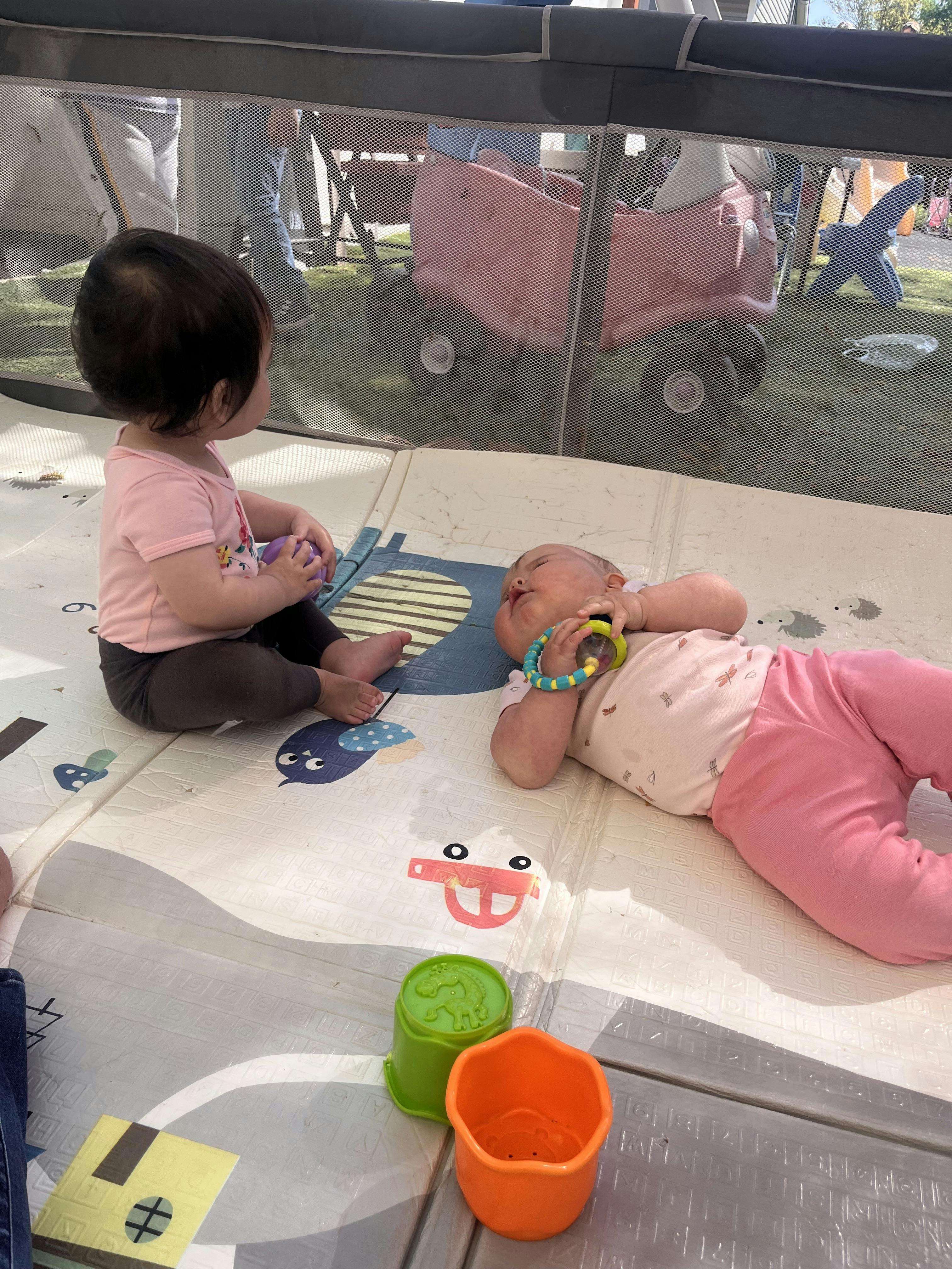 Two children sitting and lying on a play mat inside a playpen