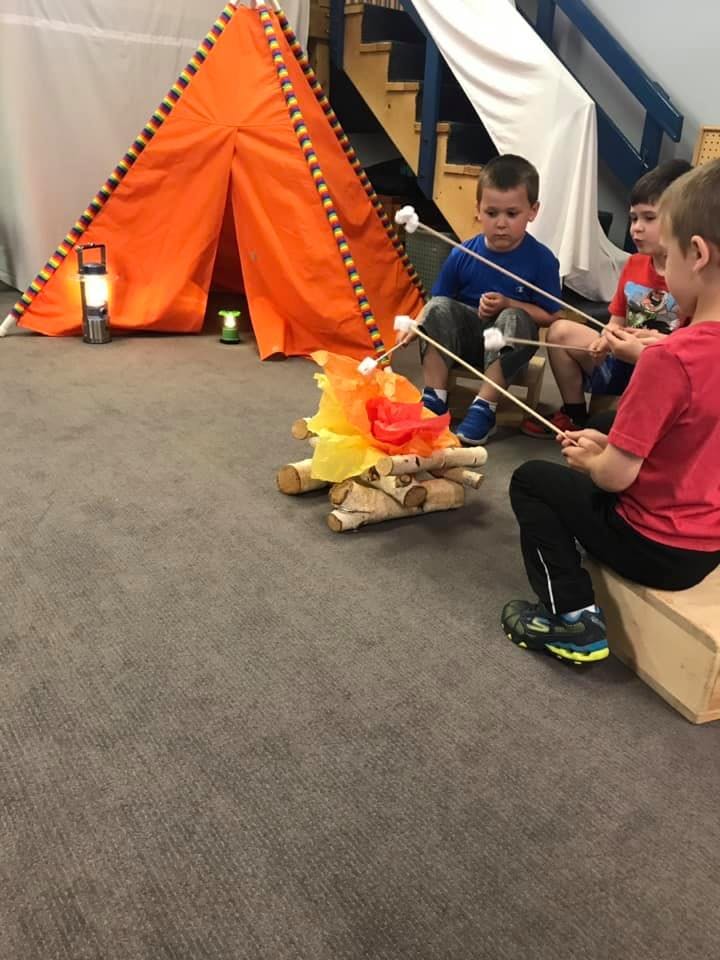 Children roasting marshmallows over a pretend campfire next to an orange tent