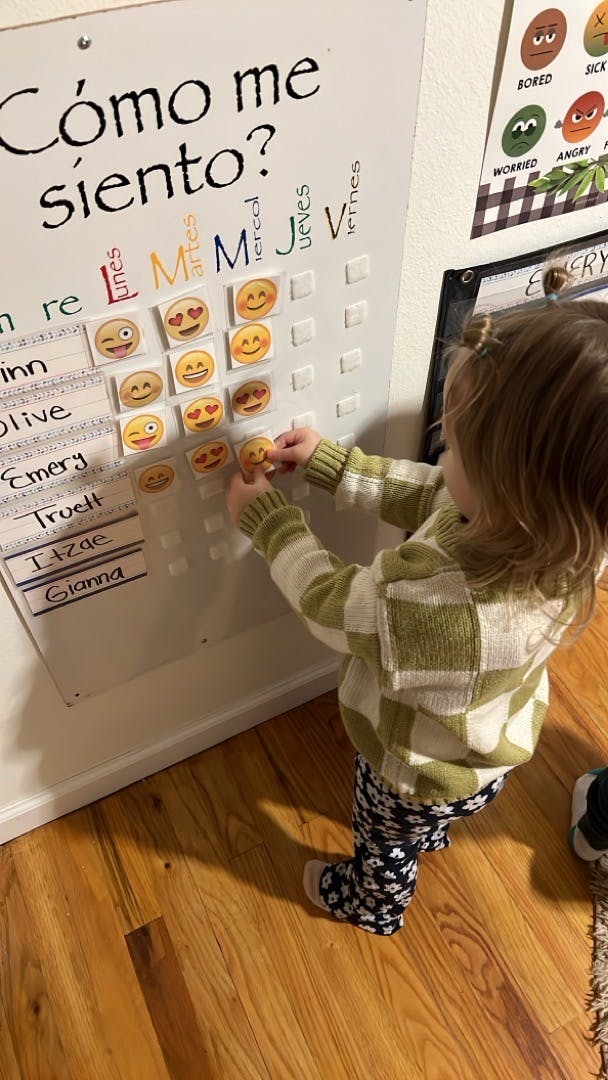 Child placing an emoji sticker on a whiteboard with "Cómo me siento?" written on it