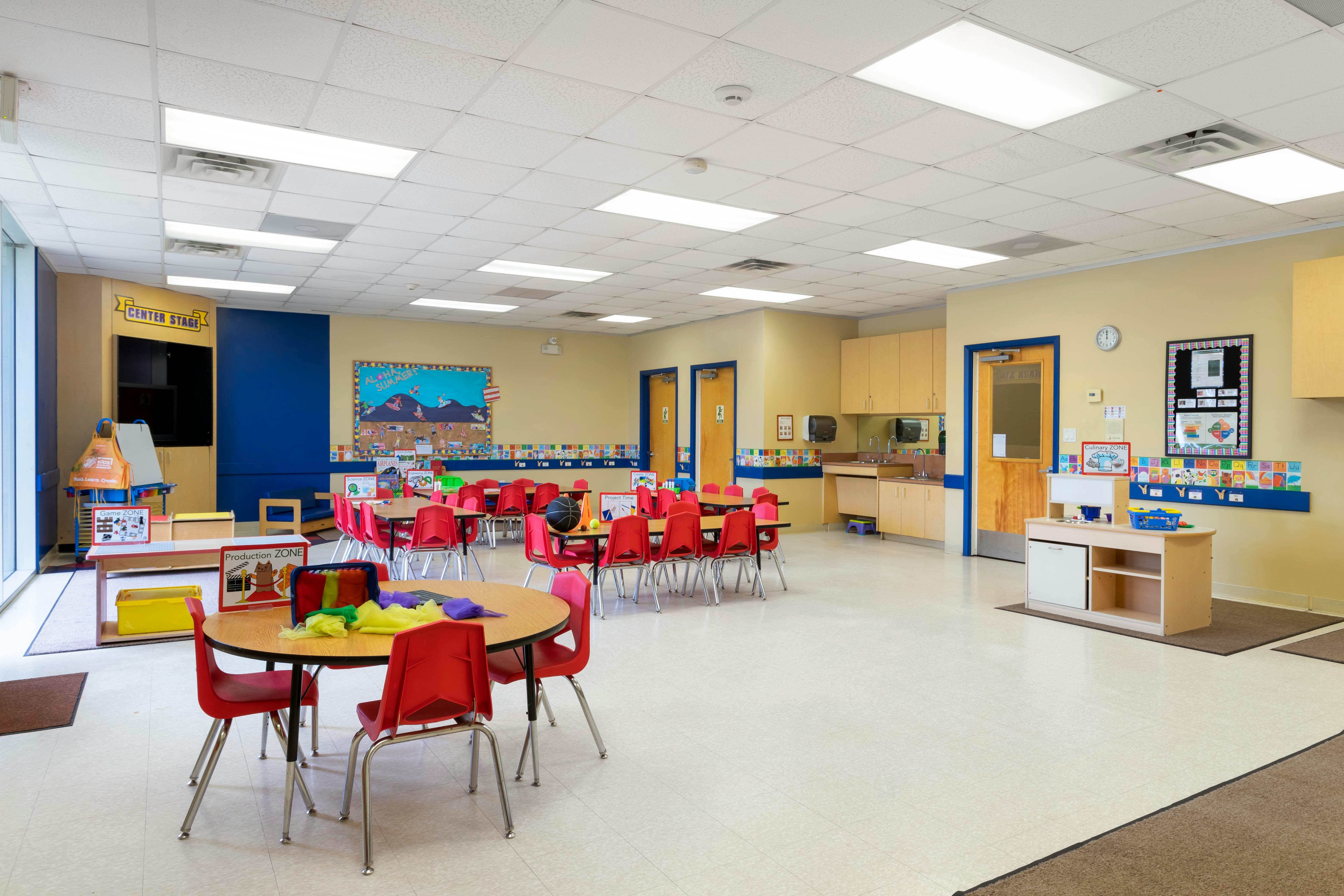 Empty classroom with small round tables and red chairs, a stage area, and activity zones