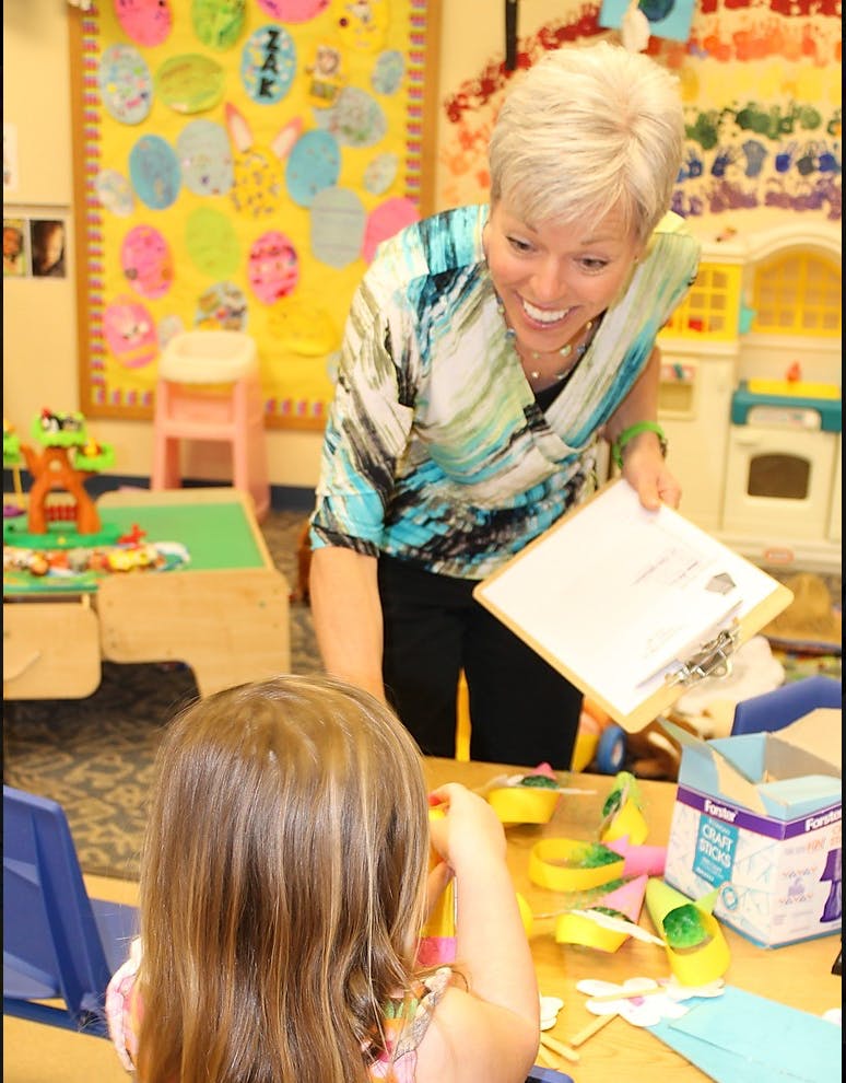 Person holding a clipboard talking to a child in a classroom