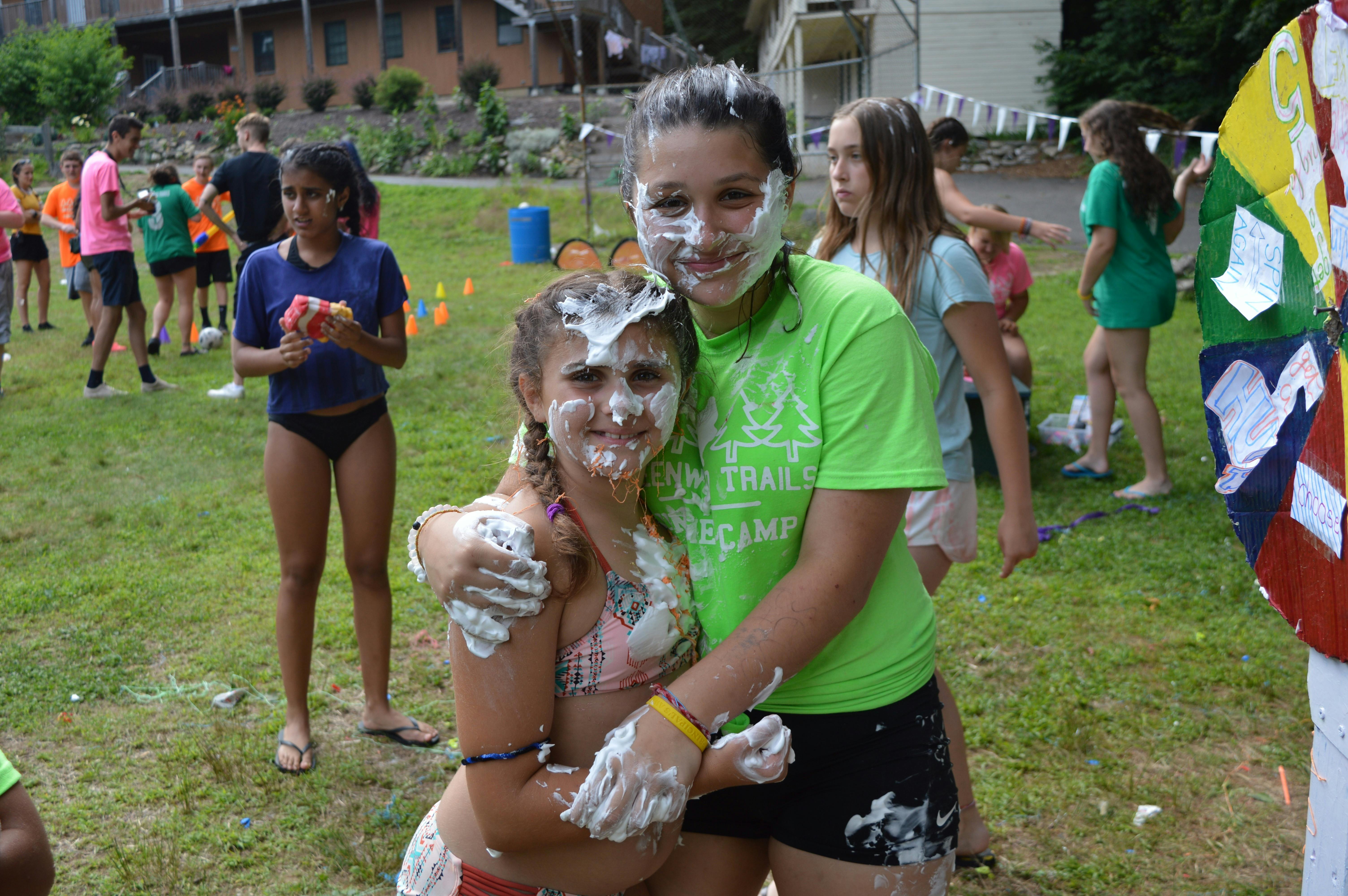 Two girls covered in white cream hugging, other children playing in background
