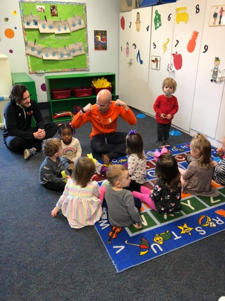 Two adults and multiple children sitting on a rug in a classroom