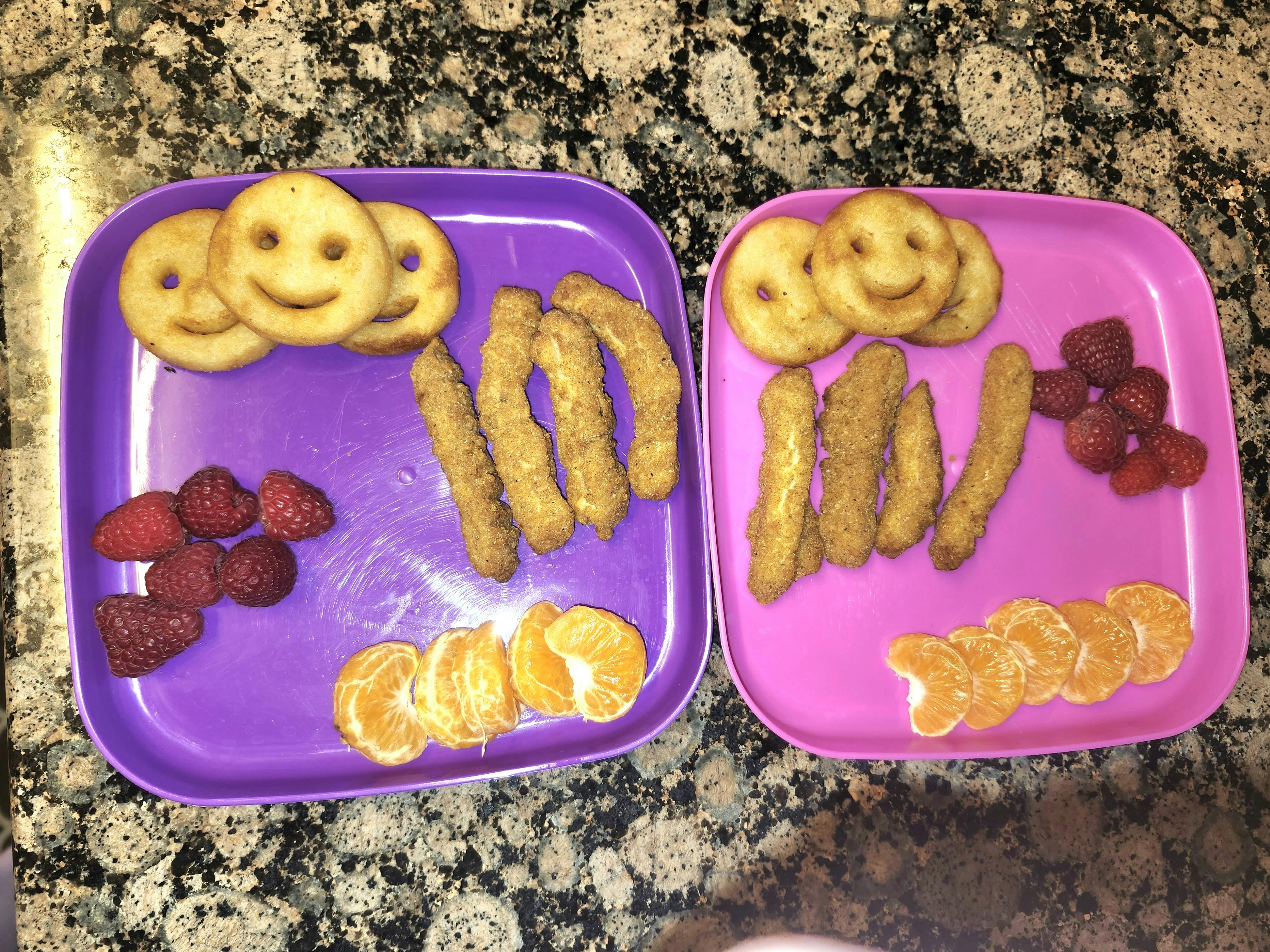 Two plates of food with smiley face fries, chicken strips, raspberries, and orange slices