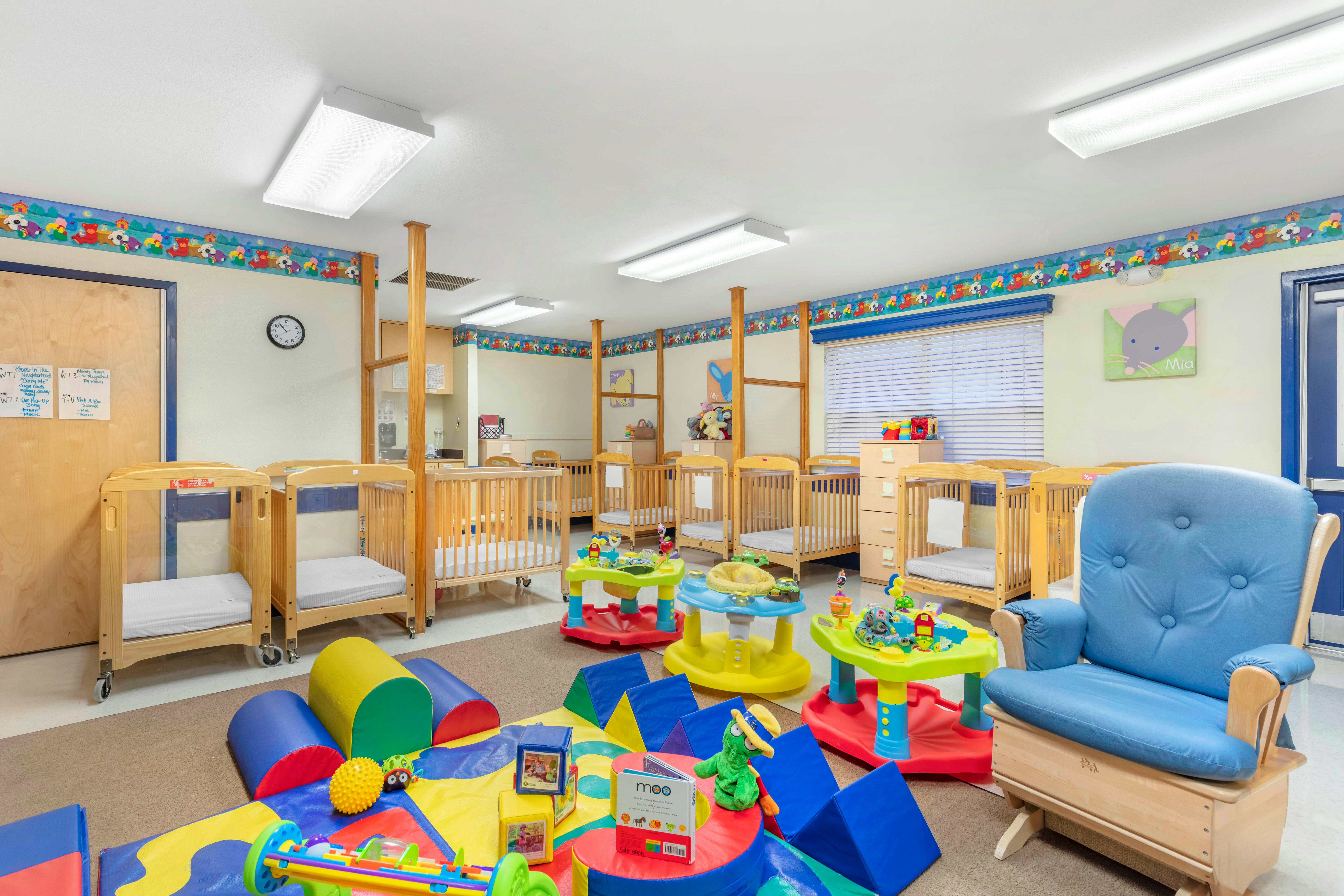 A colorful infant room with multiple cribs, a blue rocking chair, and play mats