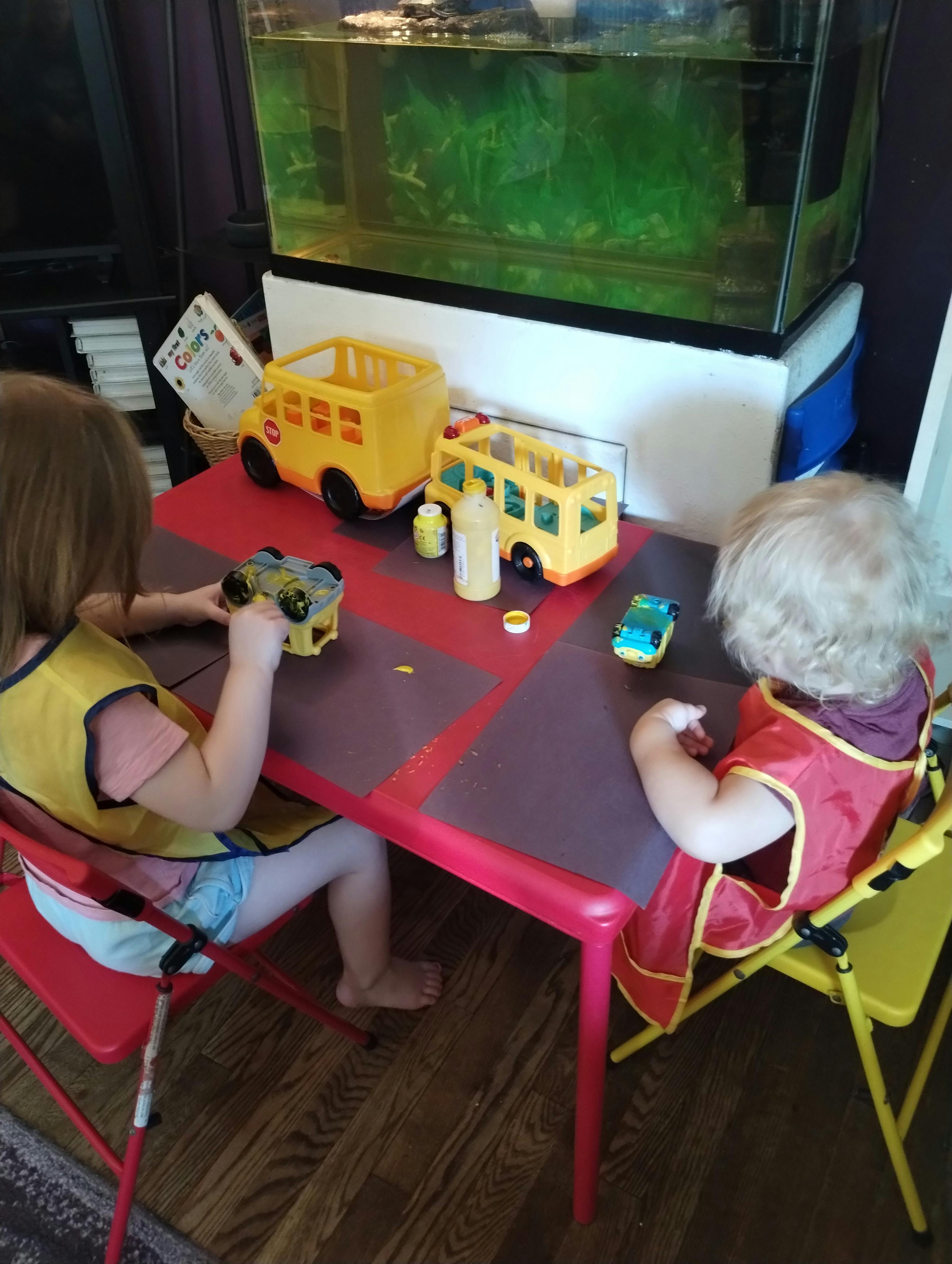 Two children wearing art smocks playing with toy vehicles at a red table