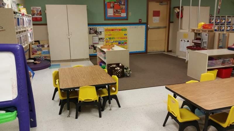 An empty classroom with small tables and chairs, shelves, and a bulletin board
