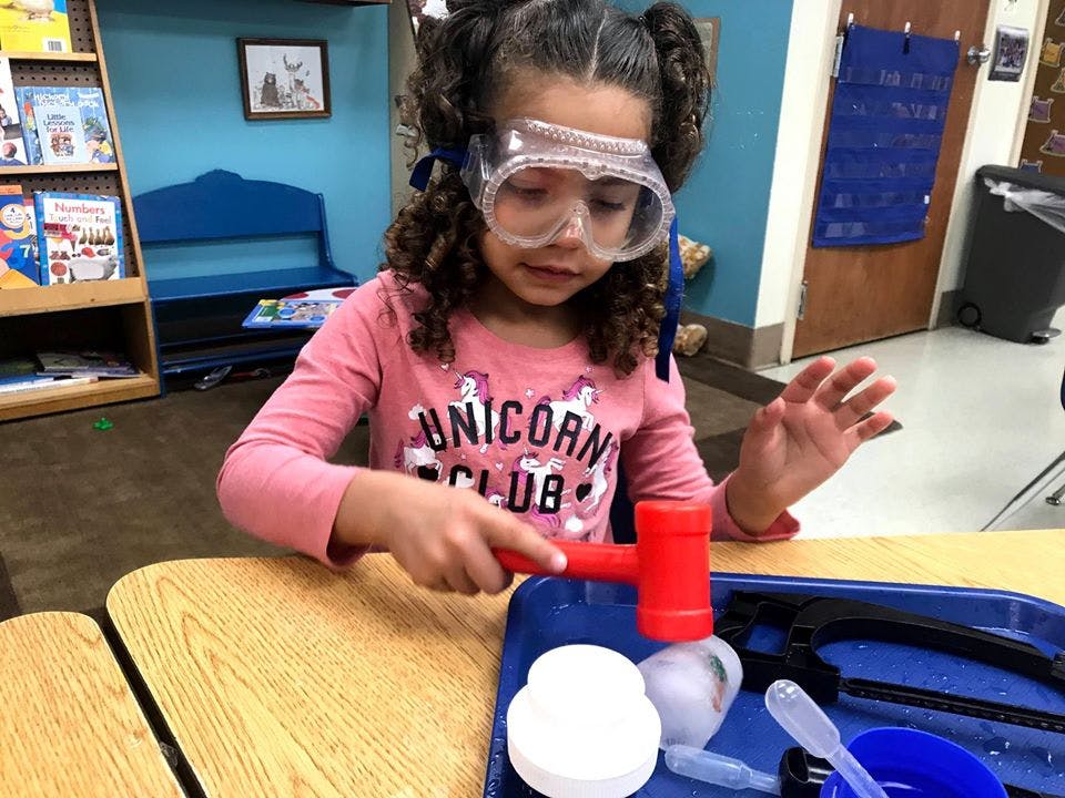 Child wearing safety goggles and holding a red hammer over a tray of tray of items