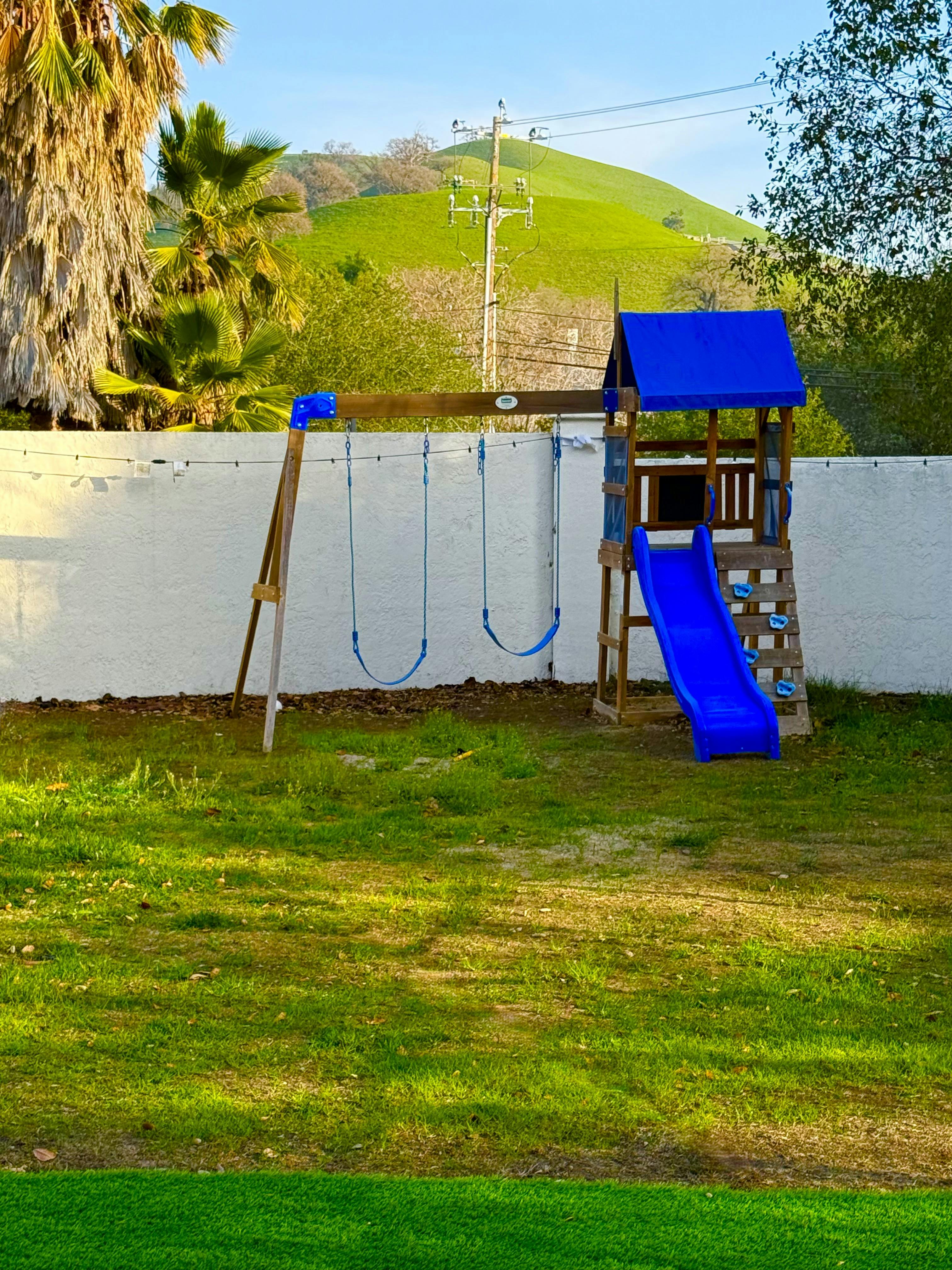 Wooden playset with two swings, a blue slide, and a climbing wall on green grass